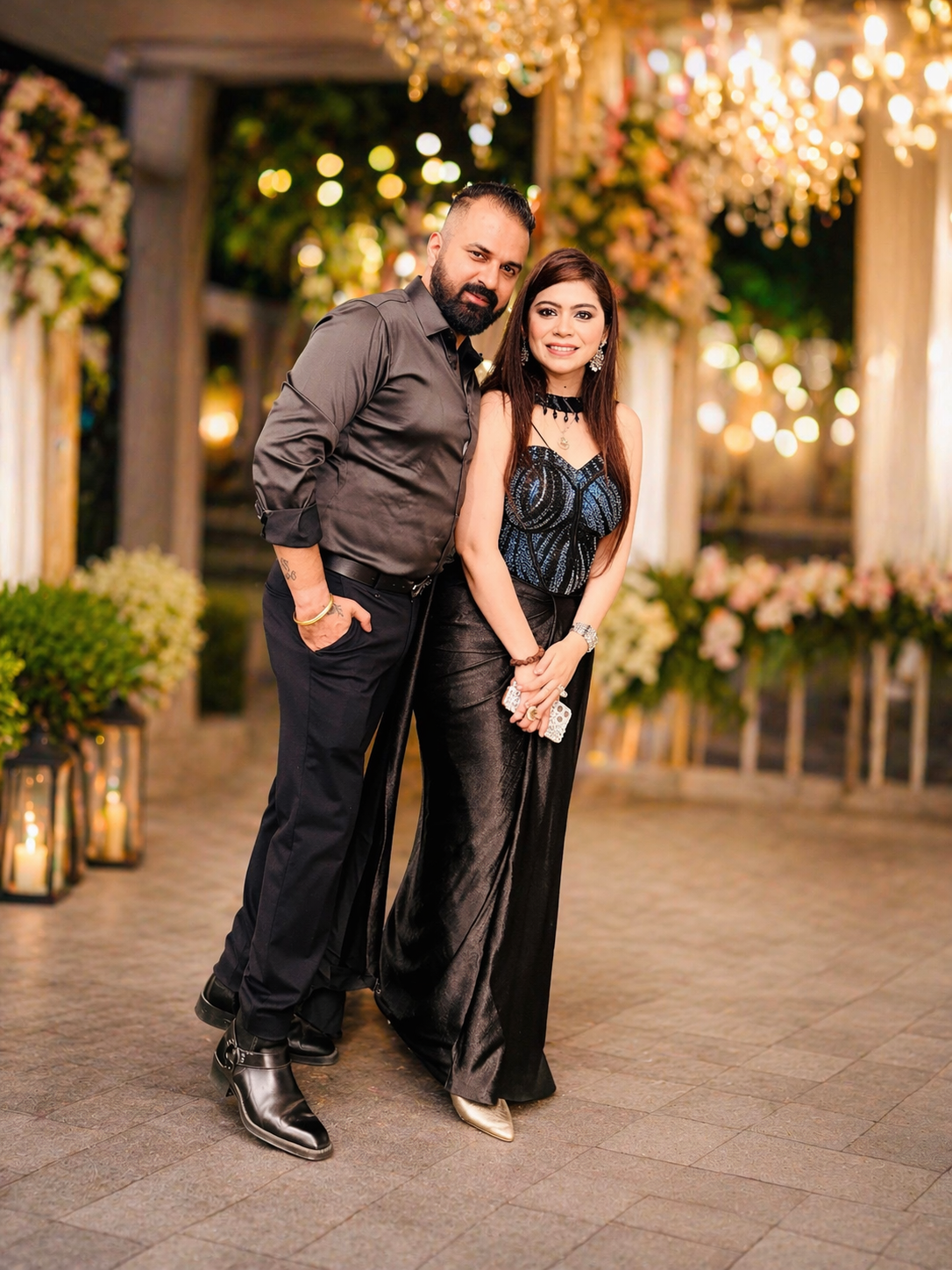 Man and woman posing together in formal attire at an elegant outdoor event with floral decorations and lights.